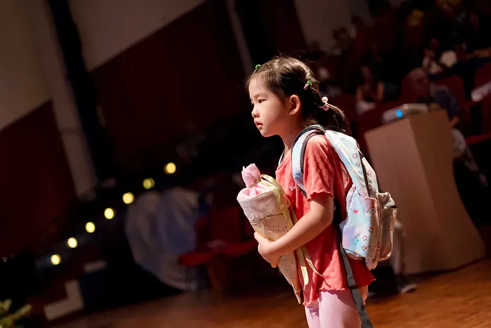 A little girl with a backpack is standing on a stage holding a stuffed animal.