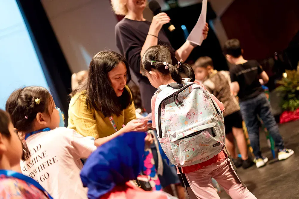 A woman is talking into a microphone while a girl with a backpack looks on.