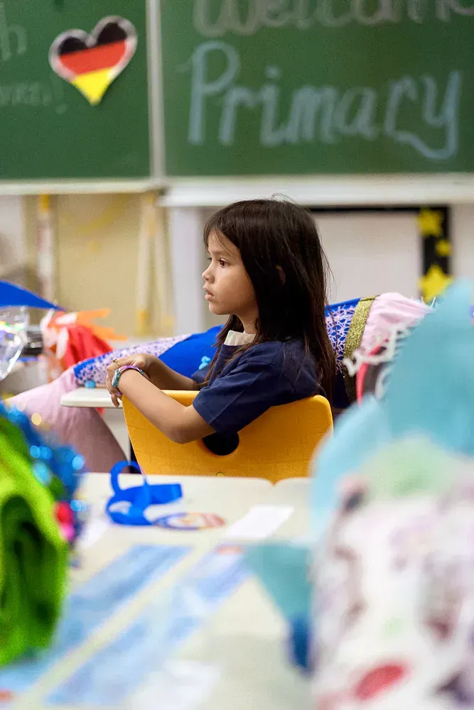 A little girl is sitting at a desk in front of a chalkboard that says primary