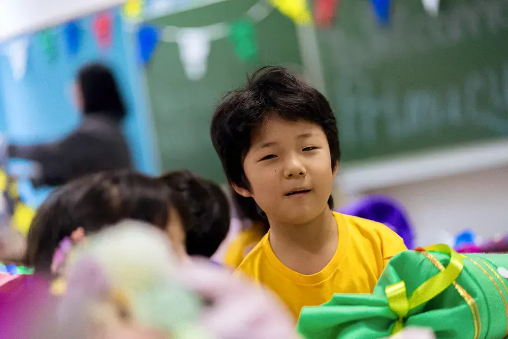 A young boy in a yellow shirt is sitting in a classroom with other children.