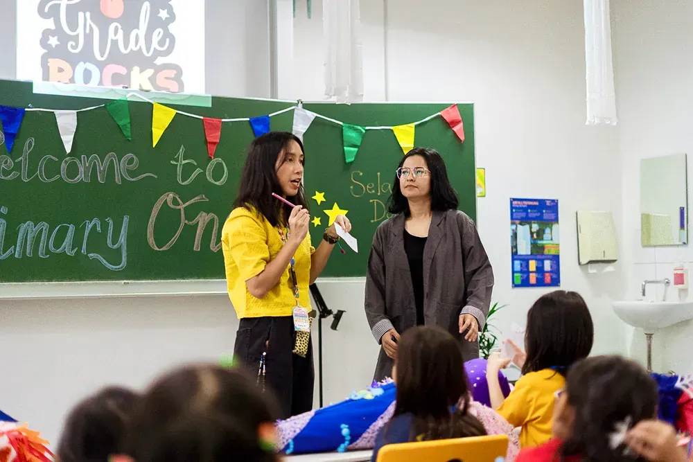 Two women are standing in front of a group of children in a classroom.