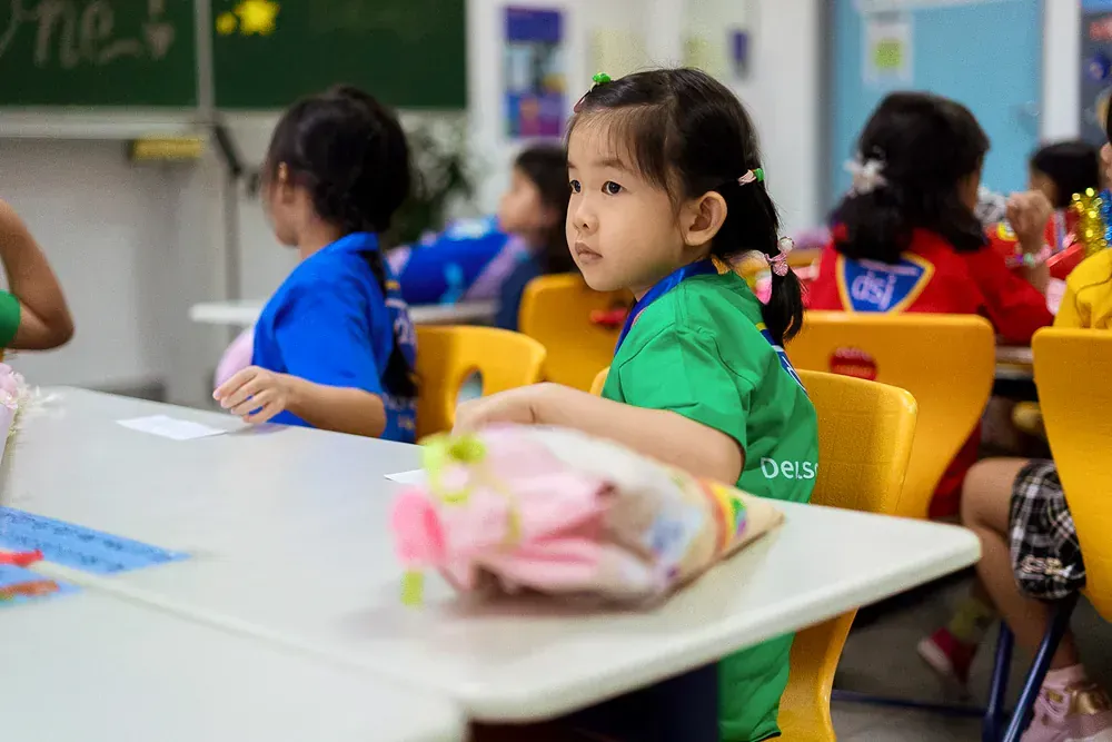 A little girl is sitting at a desk in a classroom with other children.