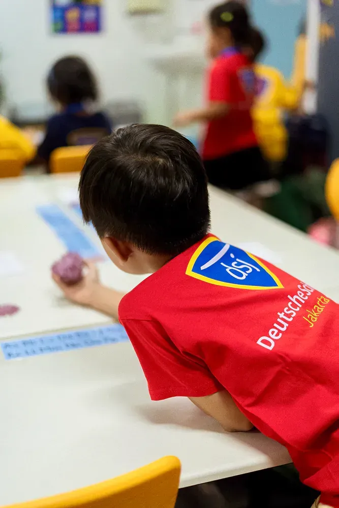 A young boy in a red shirt is sitting at a table in a classroom.