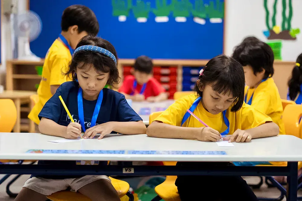 A group of children are sitting at a table in a classroom writing on a piece of paper.