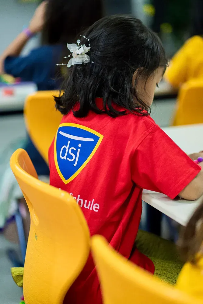 A little girl wearing a red shirt with the word dsj on it is sitting at a table.