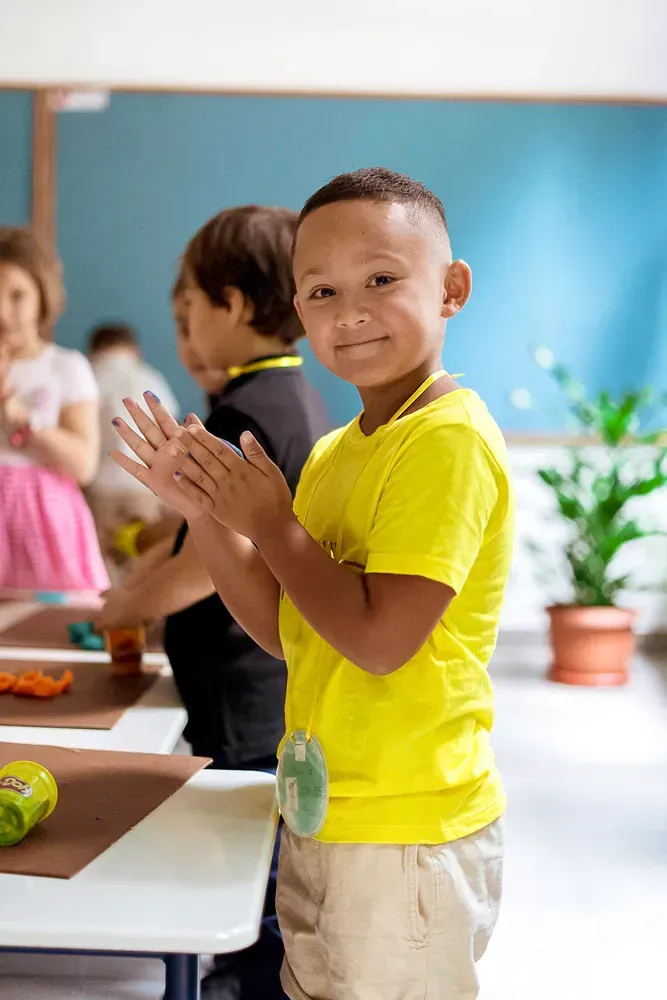 A young boy in a yellow shirt is clapping his hands in a classroom.