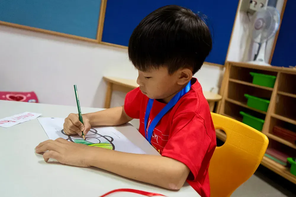 A young boy in a red shirt is sitting at a table drawing with a pencil.