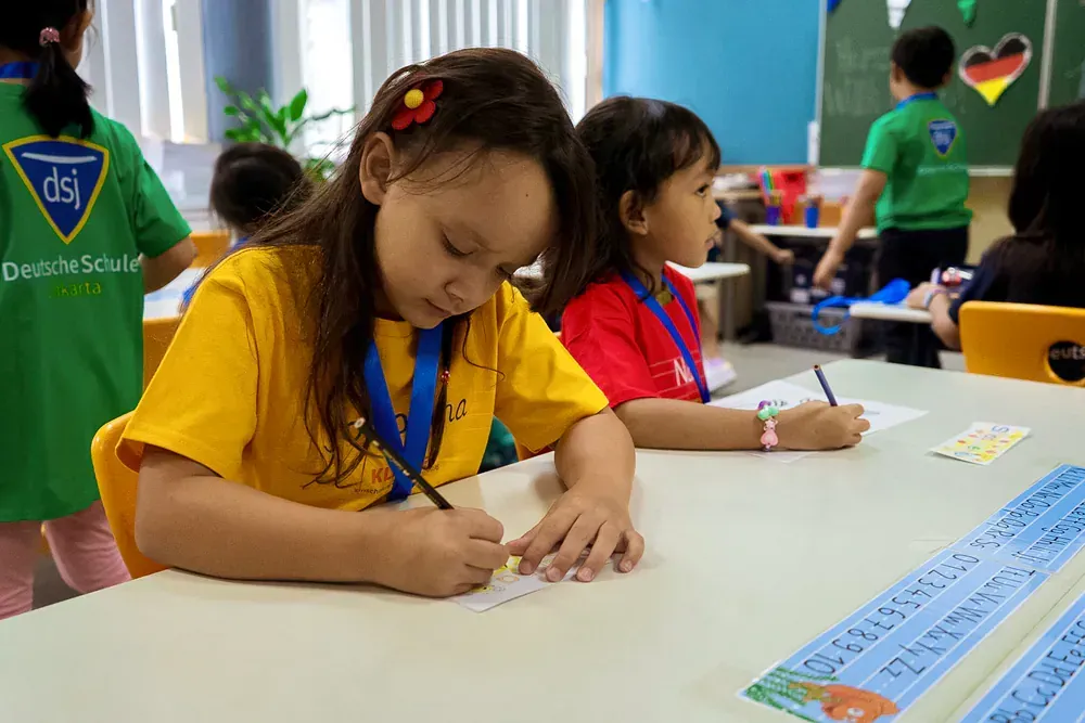 A girl in a yellow shirt is writing on a piece of paper in a classroom.