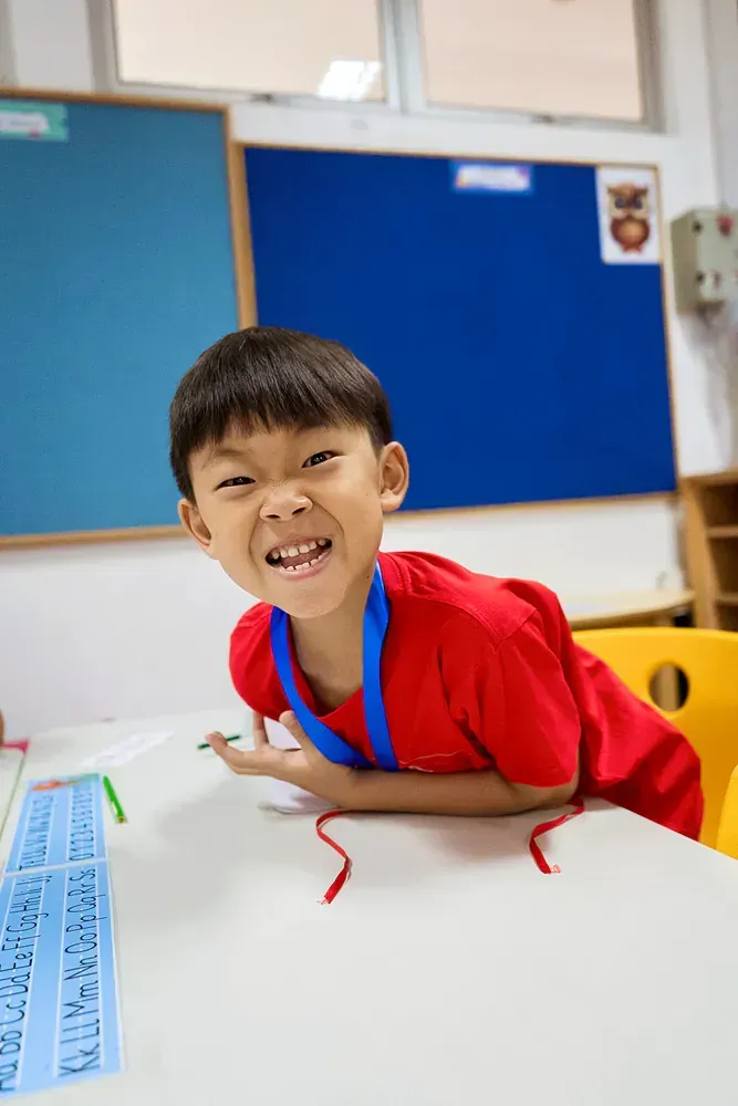A young boy in a red shirt is sitting at a table in a classroom.