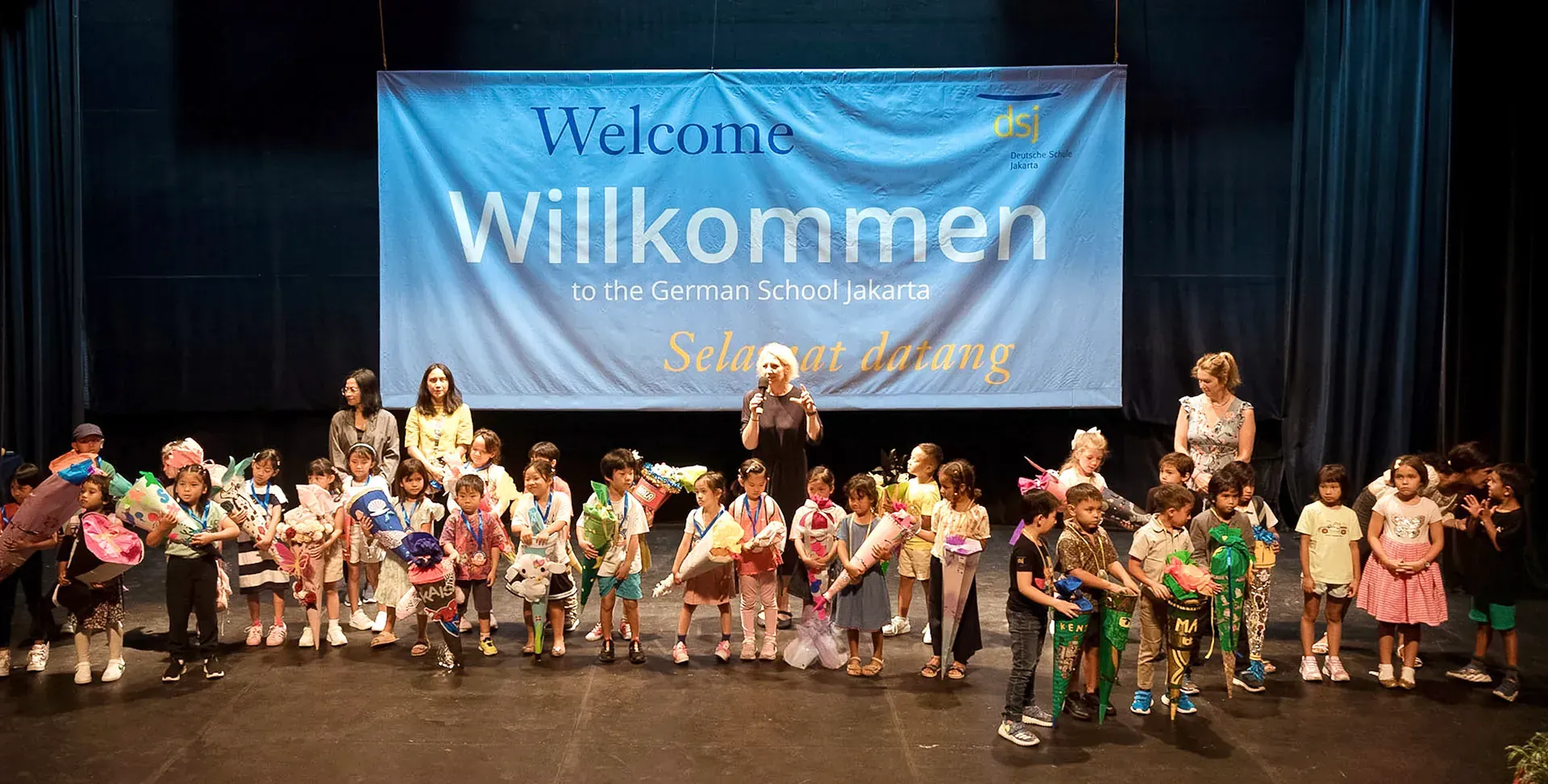 A group of children are standing on a stage in front of a welcome banner.