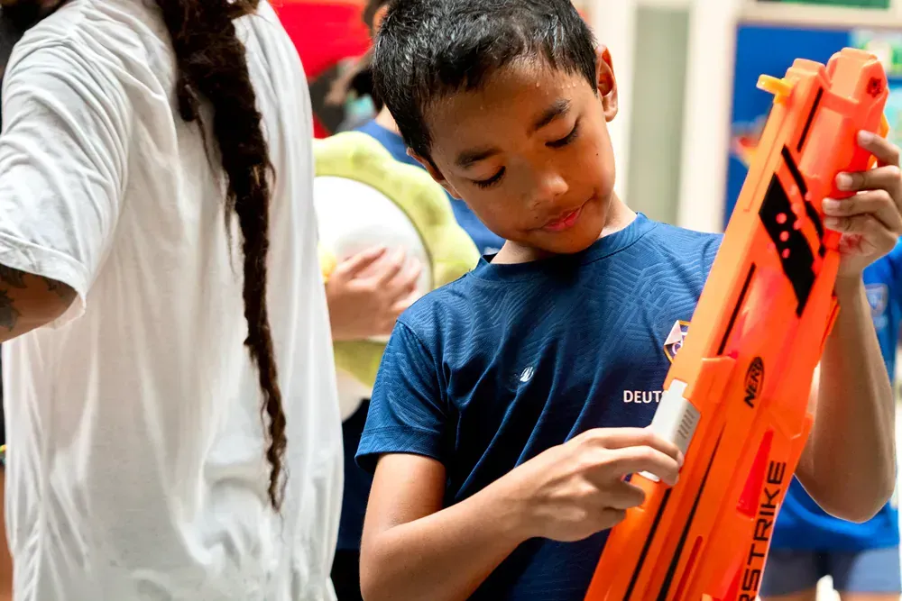 A young boy in a blue sports uniform carefully handling a bright orange toy blaster