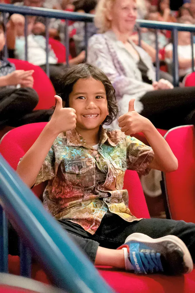 A young boy is sitting in a stadium giving a thumbs up.