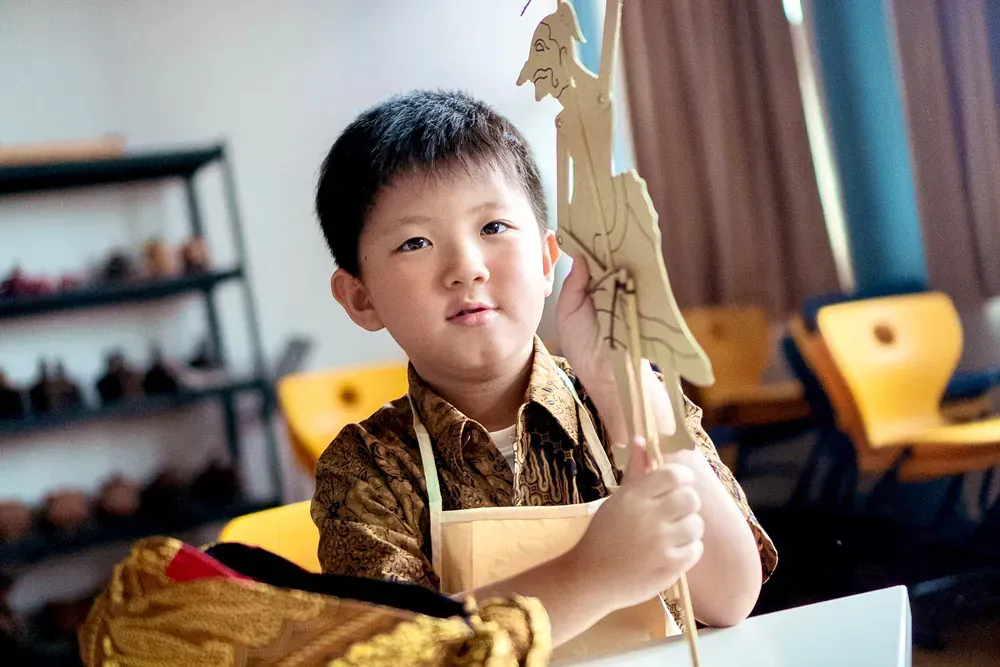 A young boy is sitting at a table holding a wooden puppet.