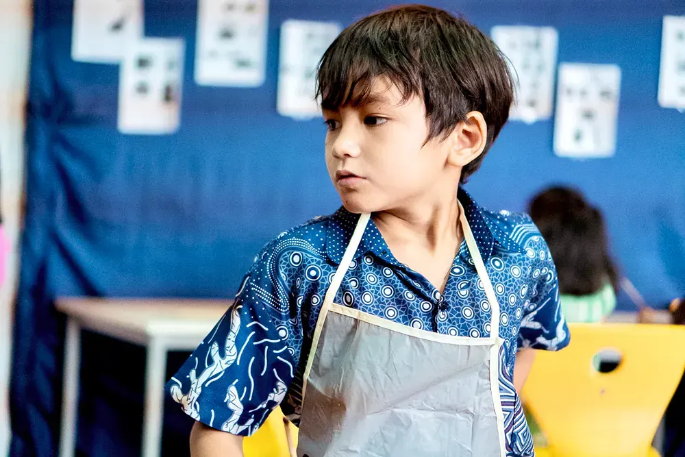 A young boy wearing an apron is standing in a classroom.