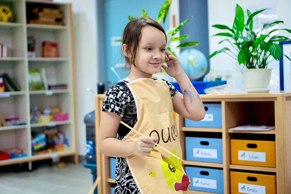 A little girl is wearing an apron and holding a brush in a classroom.