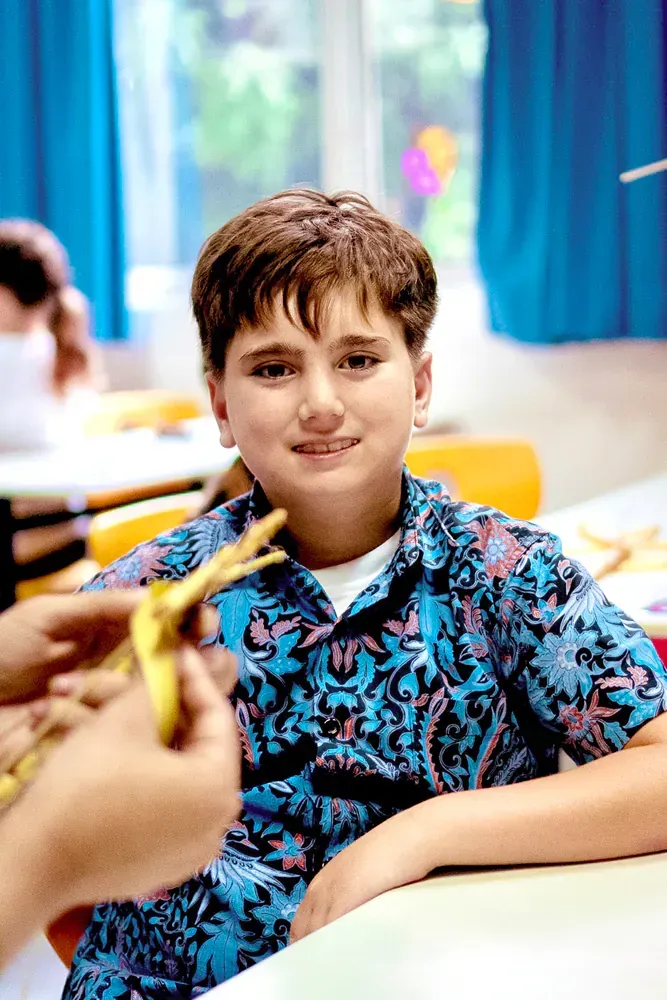 A young boy is sitting at a table in a classroom holding a banana peel.