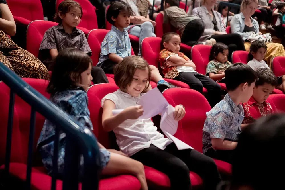 A group of children are sitting in red seats in a theater.