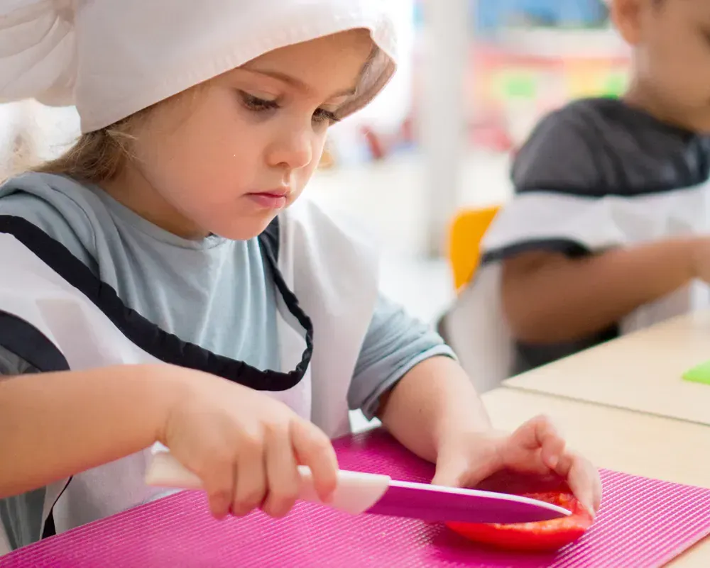Young child wearing a chef's hat and apron, carefully cutting a red vegetable with a child-safe knife on a pink cutting mat.