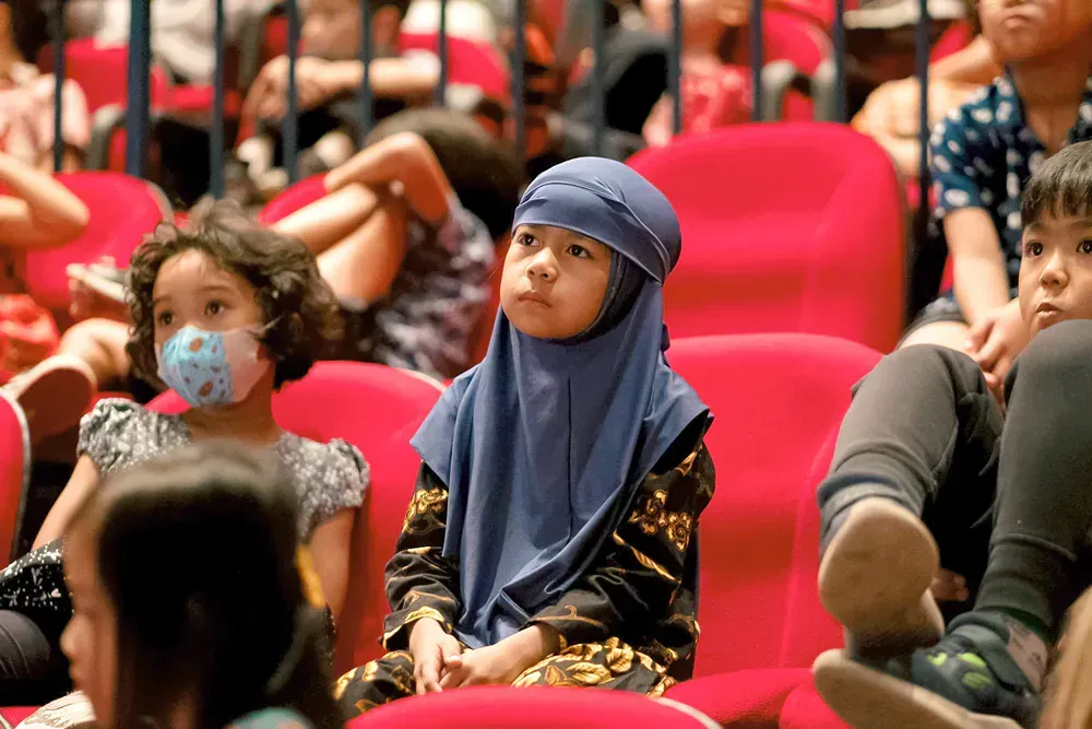 A little girl wearing a mask is sitting in a theatre.