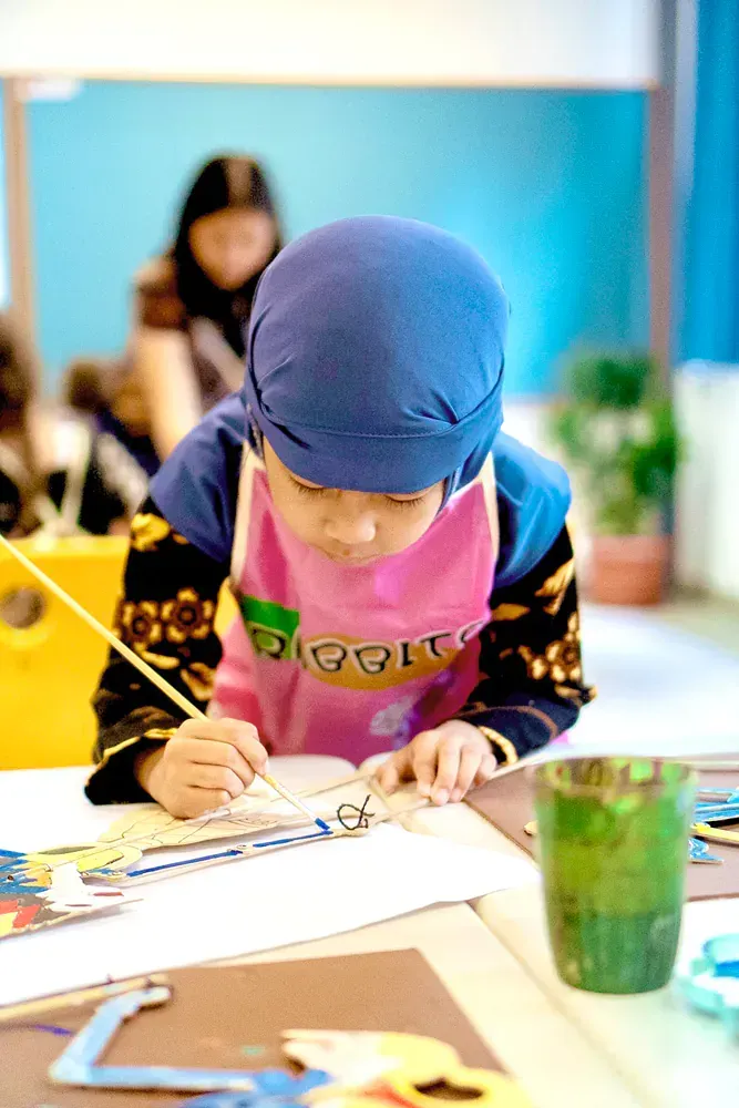 A little girl wearing a pink apron and a blue hat is painting on a piece of paper.