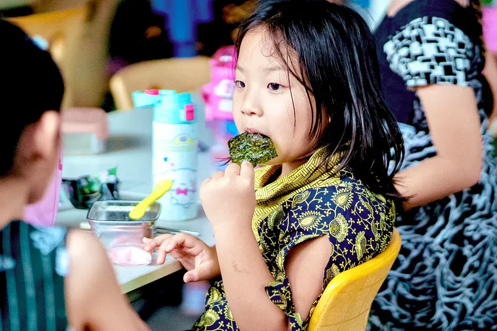 A little girl is eating a piece of broccoli while sitting at a table.