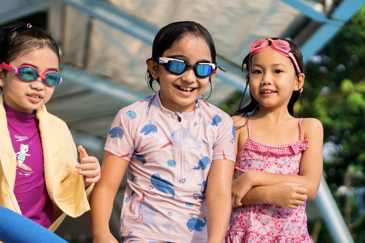 Three young girls wearing goggles are standing next to each other in front of a swimming pool.
