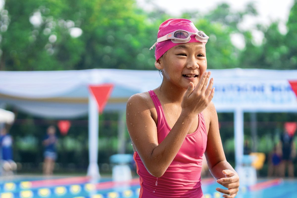 A young girl in a pink swimsuit and goggles is standing next to a swimming pool.