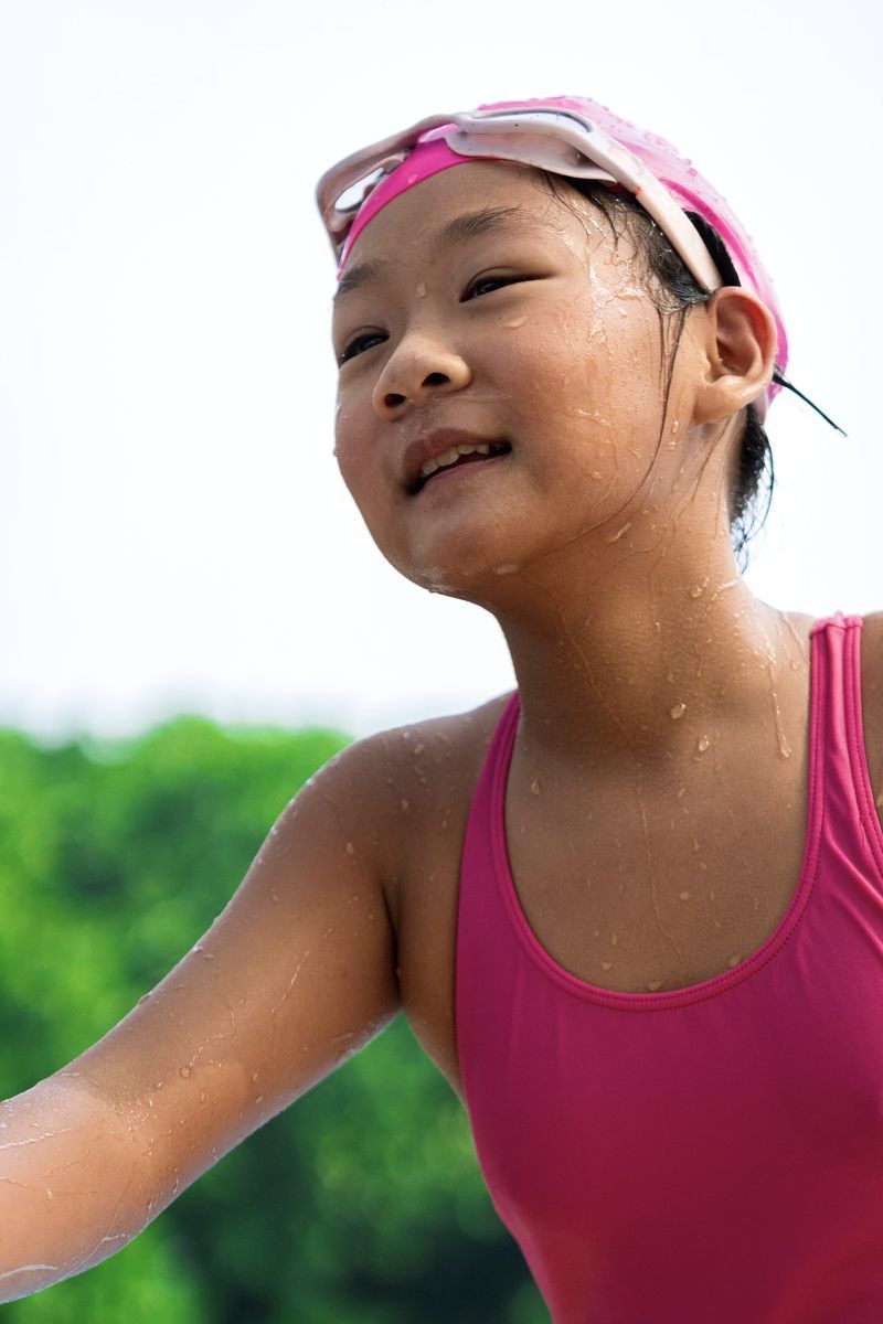A smiling young girl in a pink swimsuit and goggles appears happy and excited, possibly after a swim.