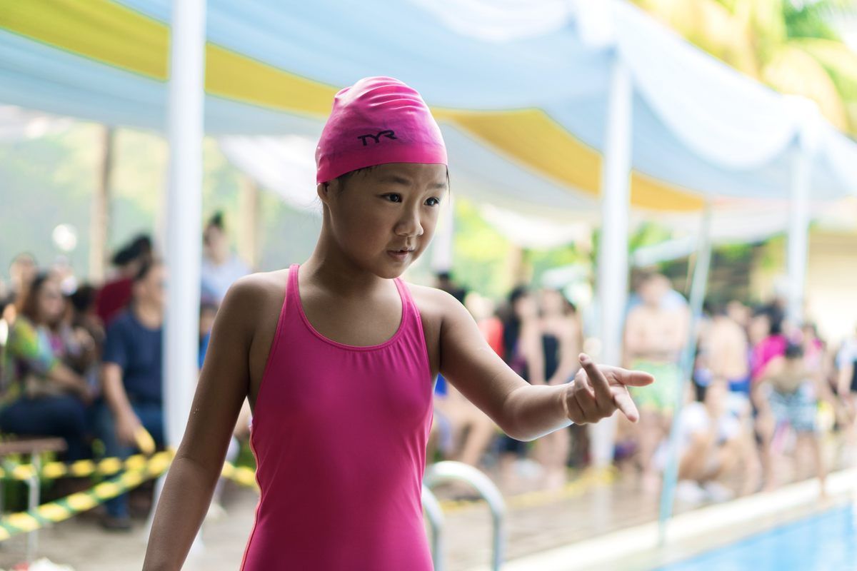 A young girl in a pink swimsuit and swim cap is standing next to a swimming pool.