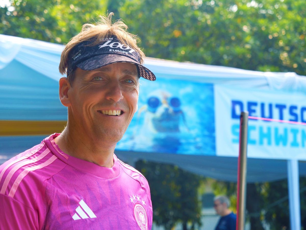 A man wearing a pink adidas shirt is smiling in front of a sign that says deutsch schwimm
