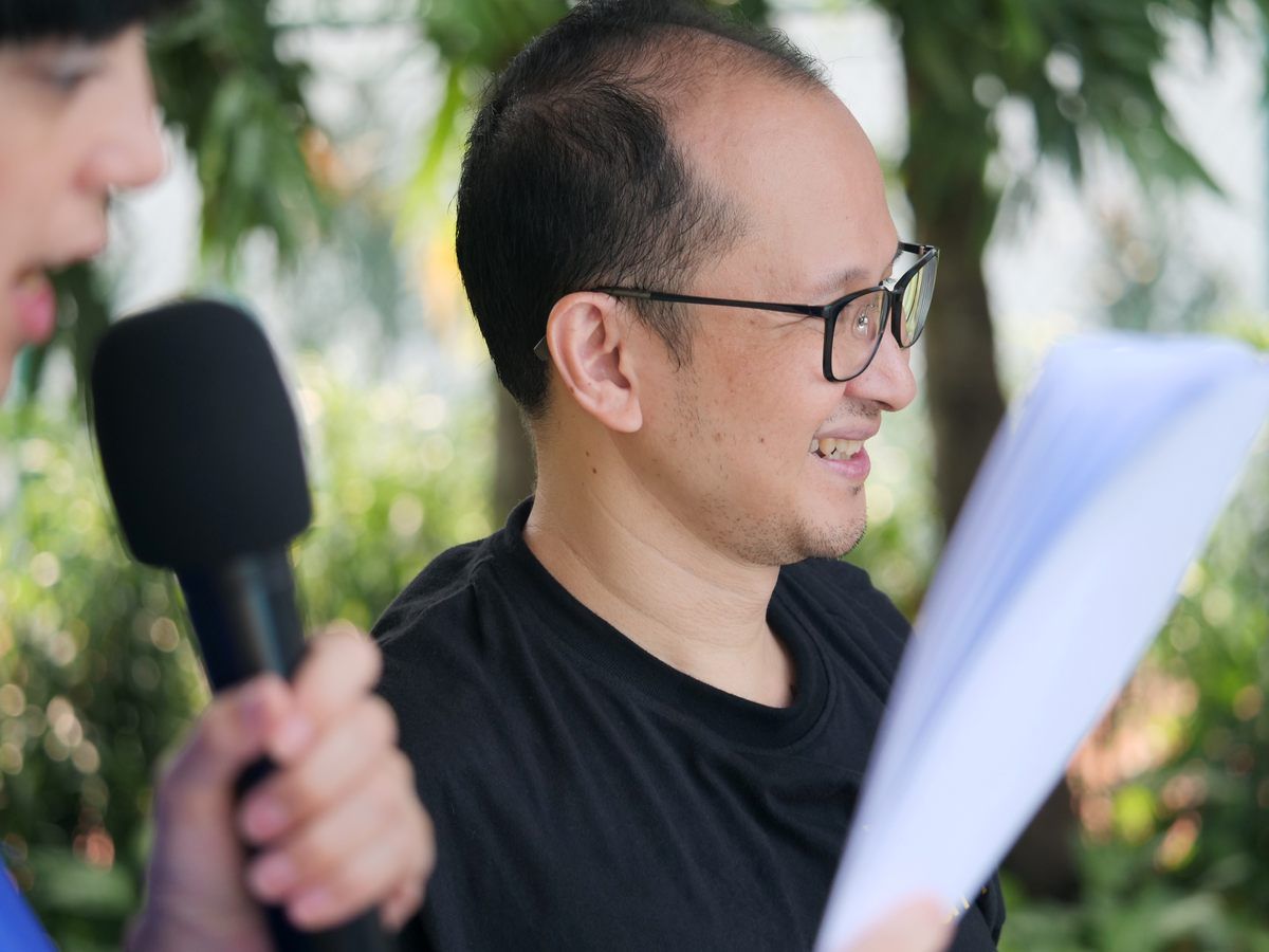 A man wearing glasses is holding a piece of paper and a microphone.