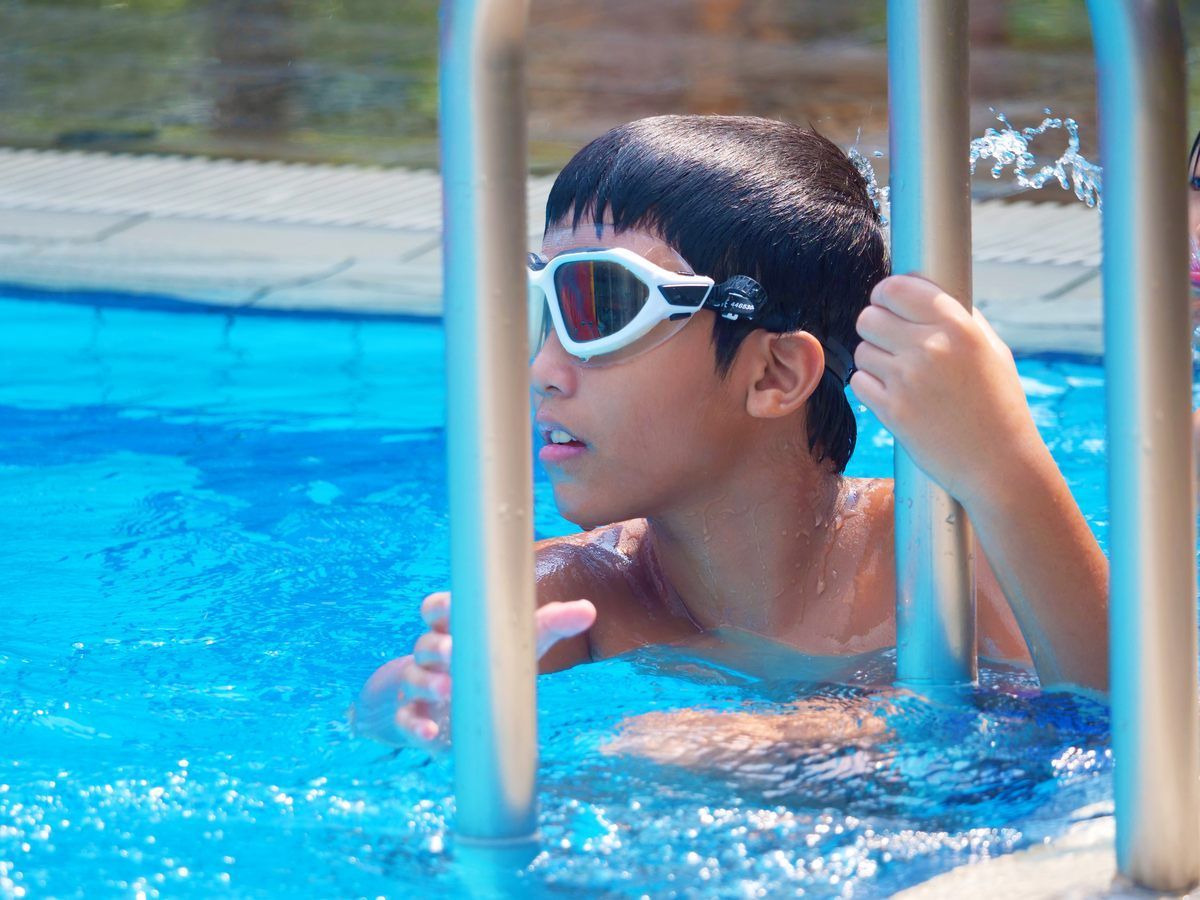 A young boy wearing goggles is getting out of a swimming pool.