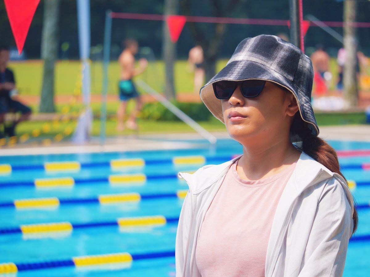 A woman wearing a hat and sunglasses is standing in front of a swimming pool.