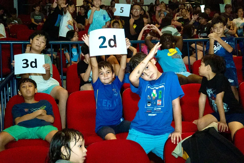 A group of children are sitting in a theater holding up signs that say 3d and 2d.