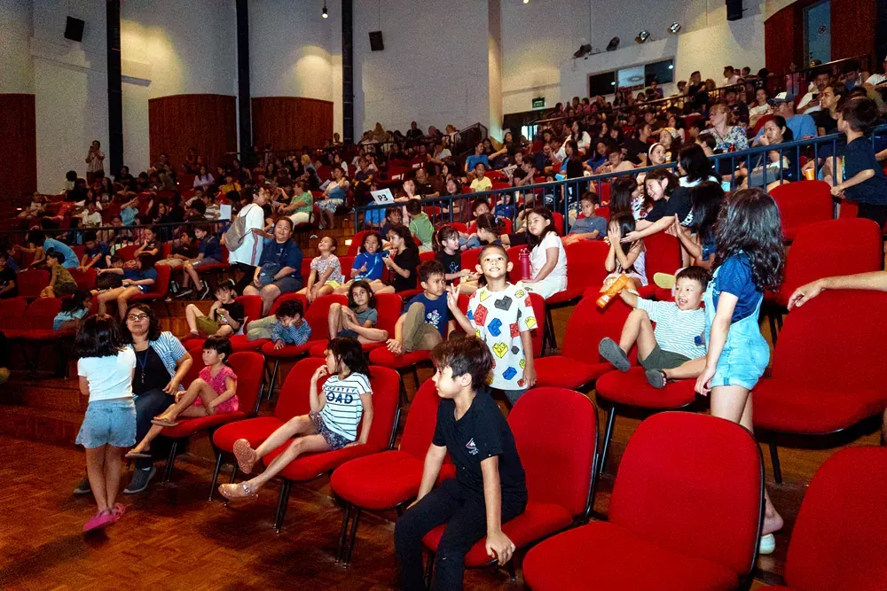 A large group of children are sitting in red chairs in an auditorium.