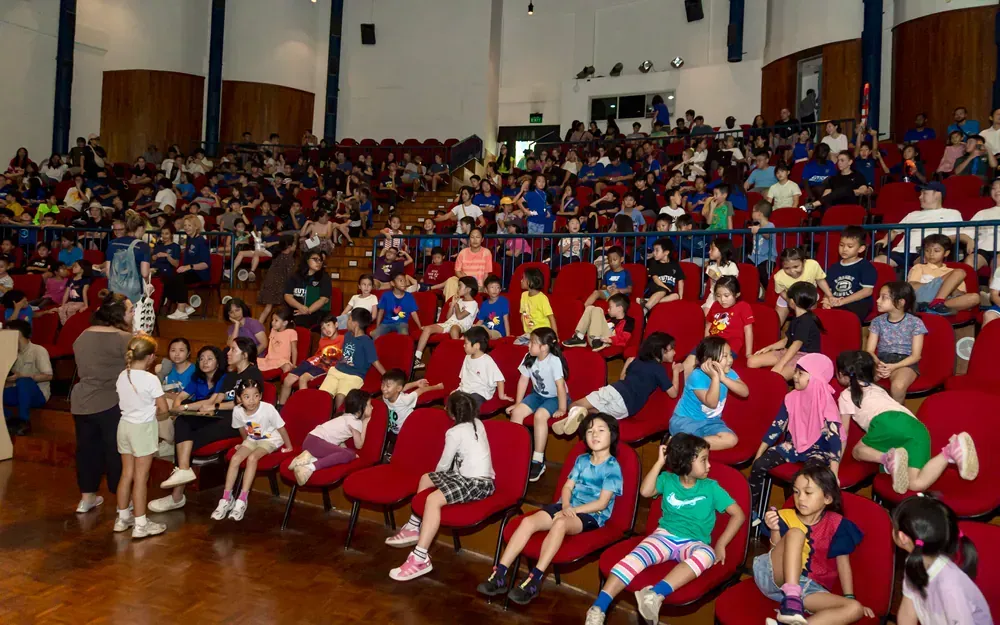 A large group of children are sitting in red chairs in an auditorium.