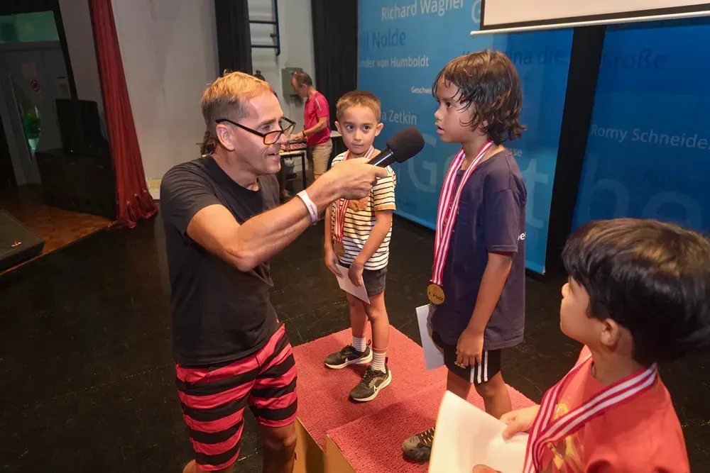A man is talking into a microphone to two young boys on a stage.
