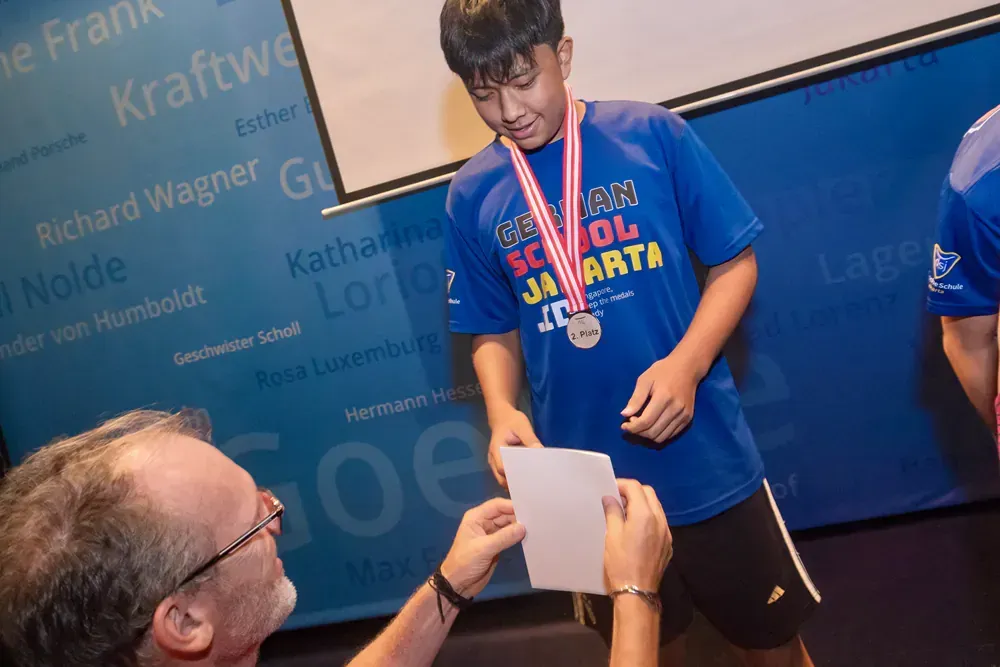 A man is giving a medal to a boy wearing a blue shirt that says school jakarta
