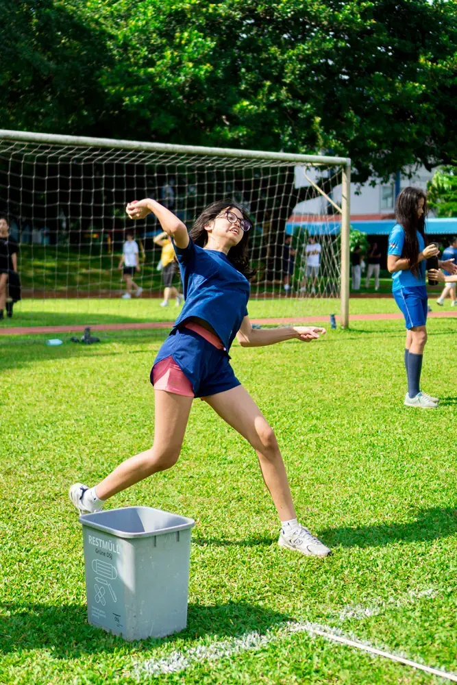 A woman is throwing a frisbee into a trash can on a soccer field.