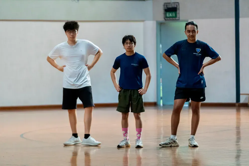 Three young men are standing in a gym with their hands on their hips.