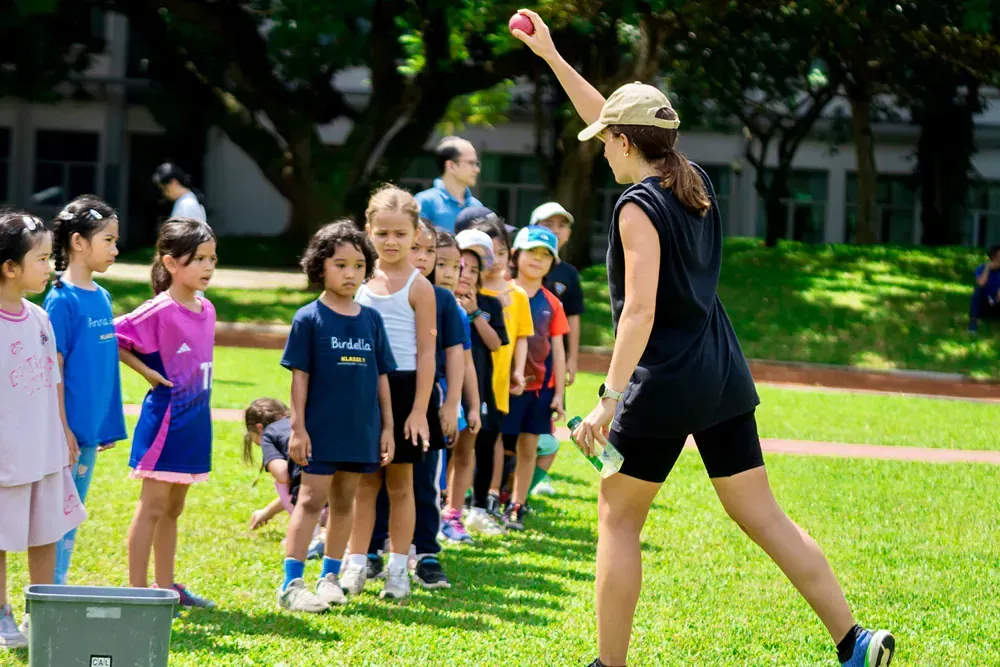 A group of children are standing in a line while a woman throws a ball.
