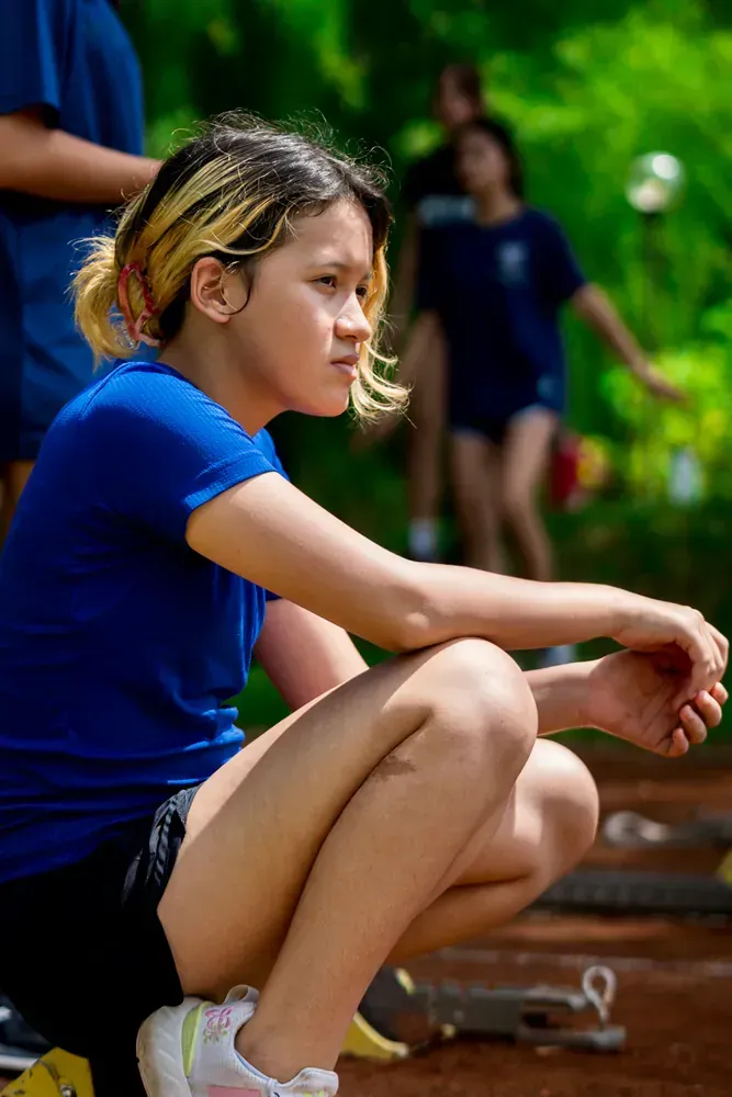 A girl in a blue sports uniform sitting on the track, watching the ongoing activities with a focused expression.