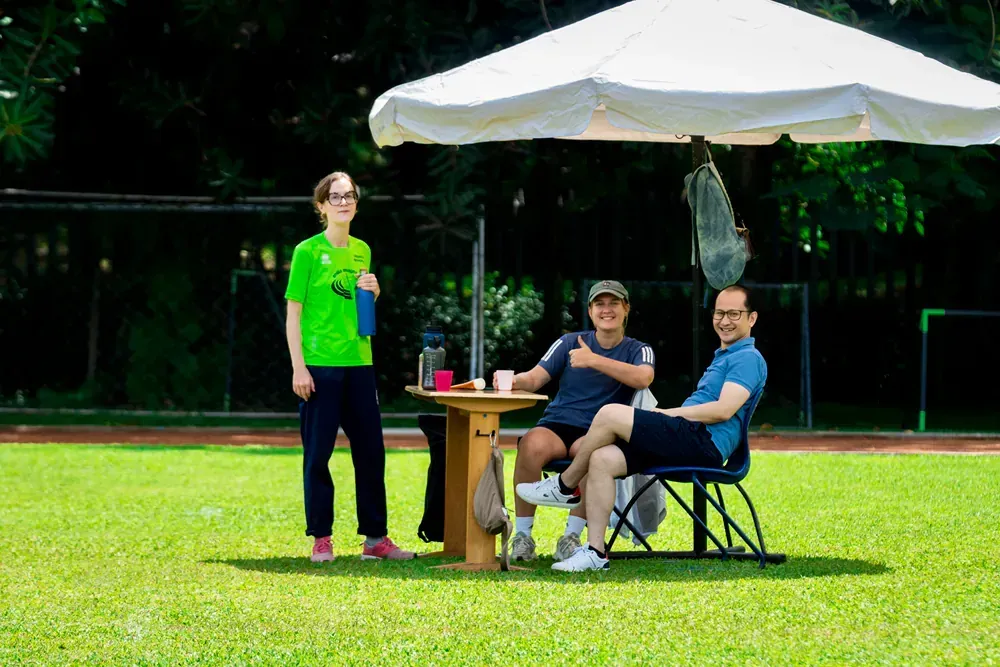 A group of people are sitting at a table under an umbrella on a field.