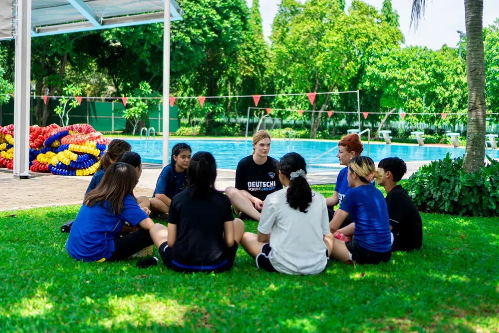 A group of people are sitting in a circle on the grass in front of a pool.