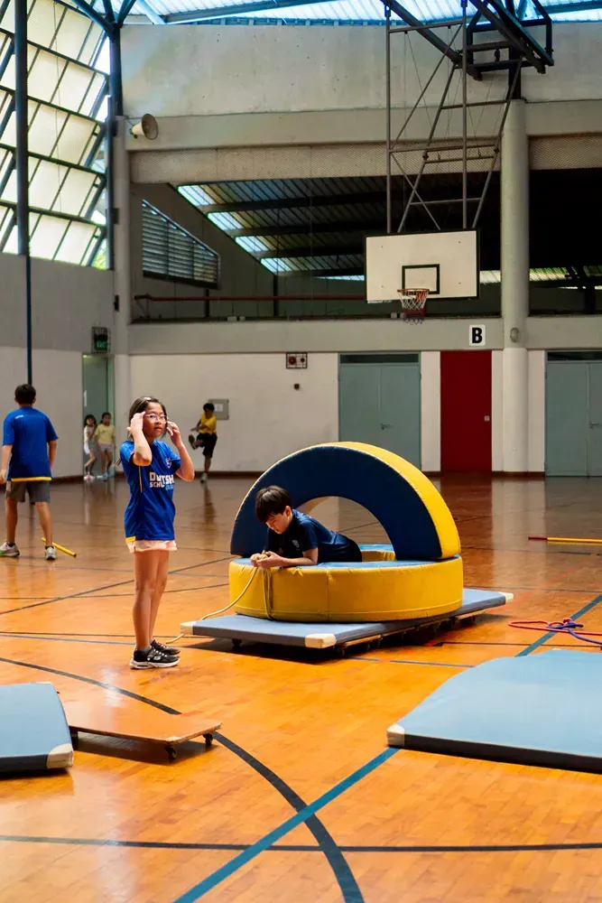 A group of children are playing in a gym with a basketball hoop in the background