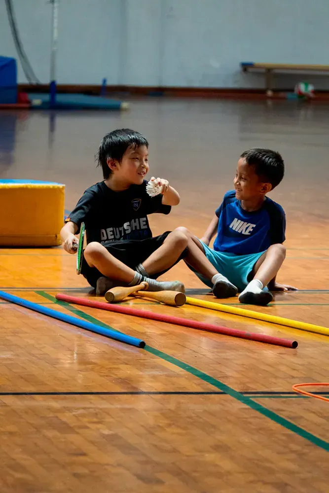 Two young boys are sitting on the floor of a gym talking to each other.