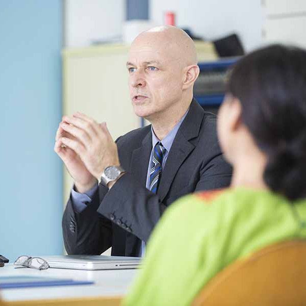 A man in a suit and tie is sitting at a table talking to a woman.