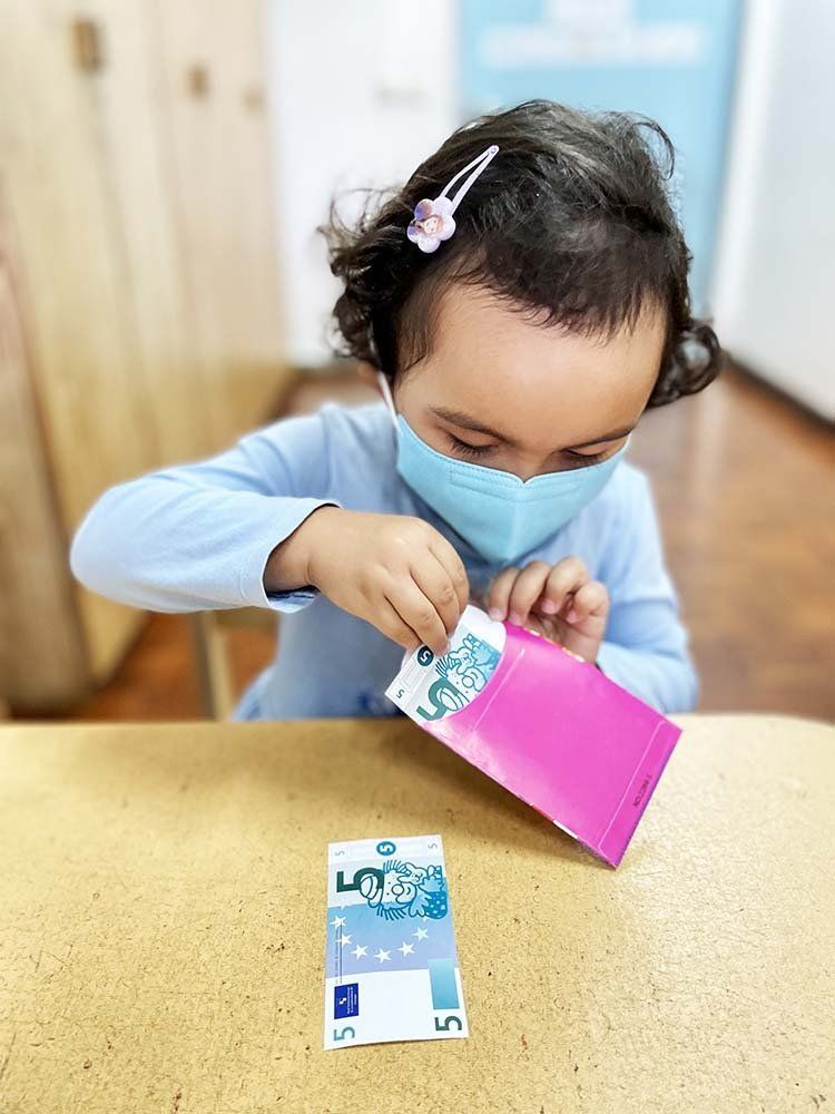 A little girl wearing a mask is putting money in a pink envelope.