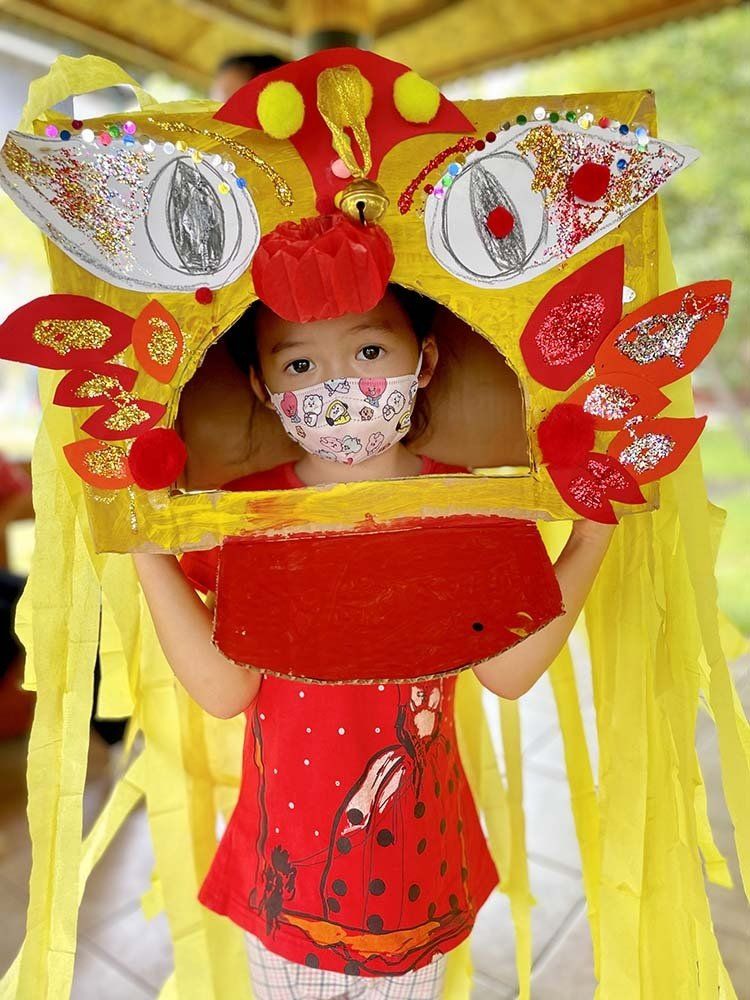 A little girl wearing a mask is holding a lion head made out of cardboard.