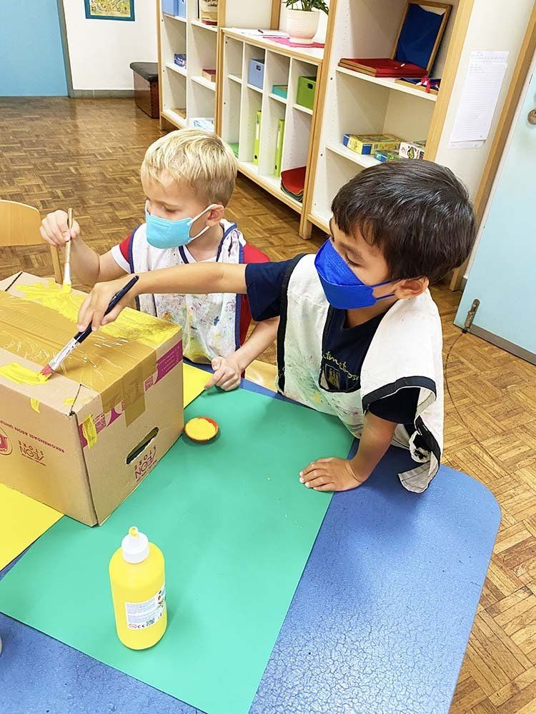 Two young boys wearing face masks are painting a cardboard box.